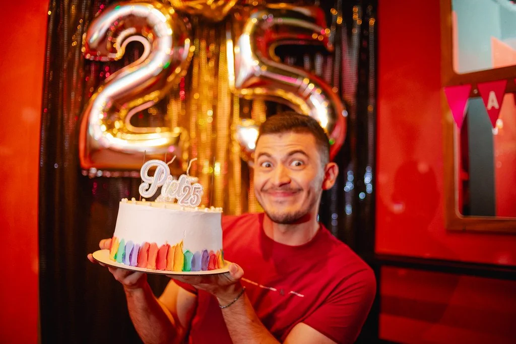 Homem sorridente segurando um bolo de aniversário branco com decoração colorida na base, com uma vela em formato 'Pi' e '25' no topo, celebrando seu 25º aniversário.
