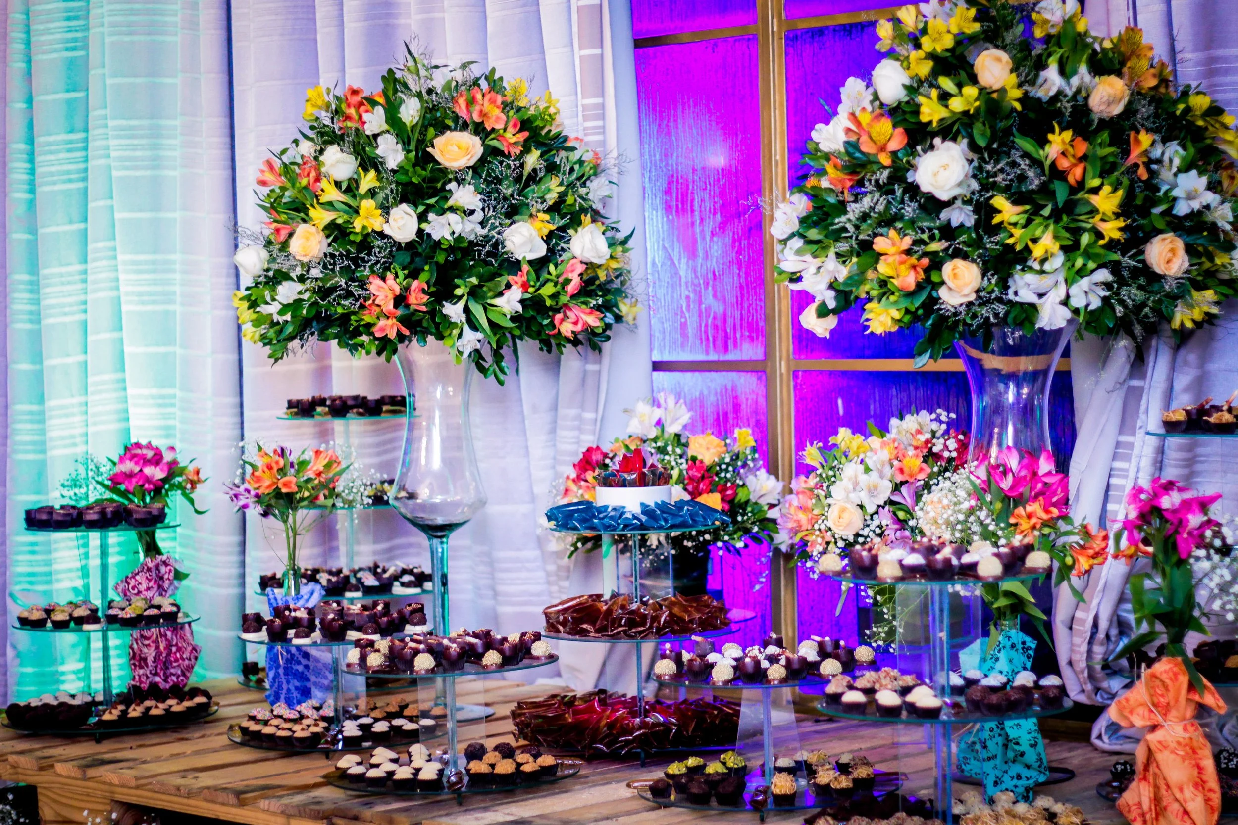 Toalha de mesa de madeira com várias bandejas de doces e flores decorativas, incluindo grandes arranjos de flores coloridas, em uma decoração de festa ou casamento.
