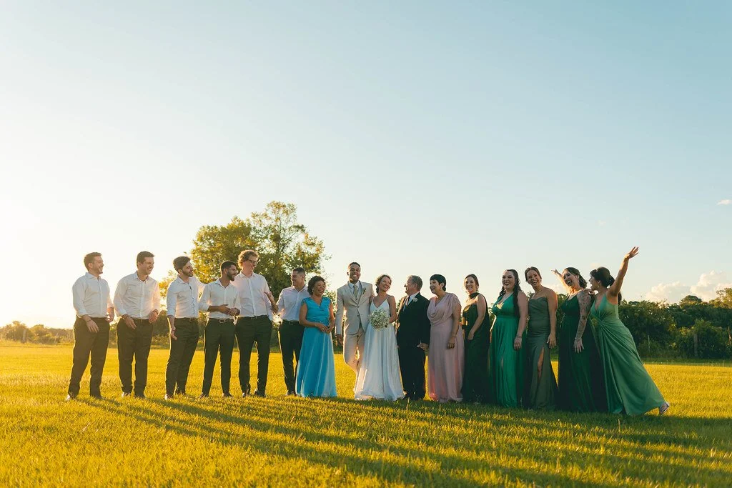 Grupo de pessoas vestindo roupas formais ao ar livre em campo com céu claro ao fundo, celebrando um casamento.