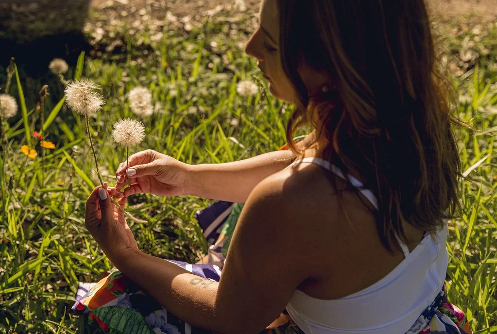 Mulher com cabelos castanhos longos segurando flores de dente-de-leão em campo ensolarado.