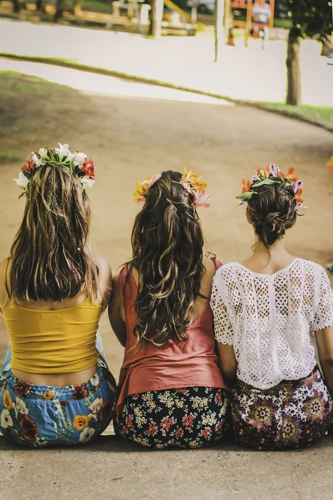 Três mulheres de costas, cada uma usando uma saia ou vestido floral e uma coroa de flores na cabeça, sentadas no chão ao ar livre.