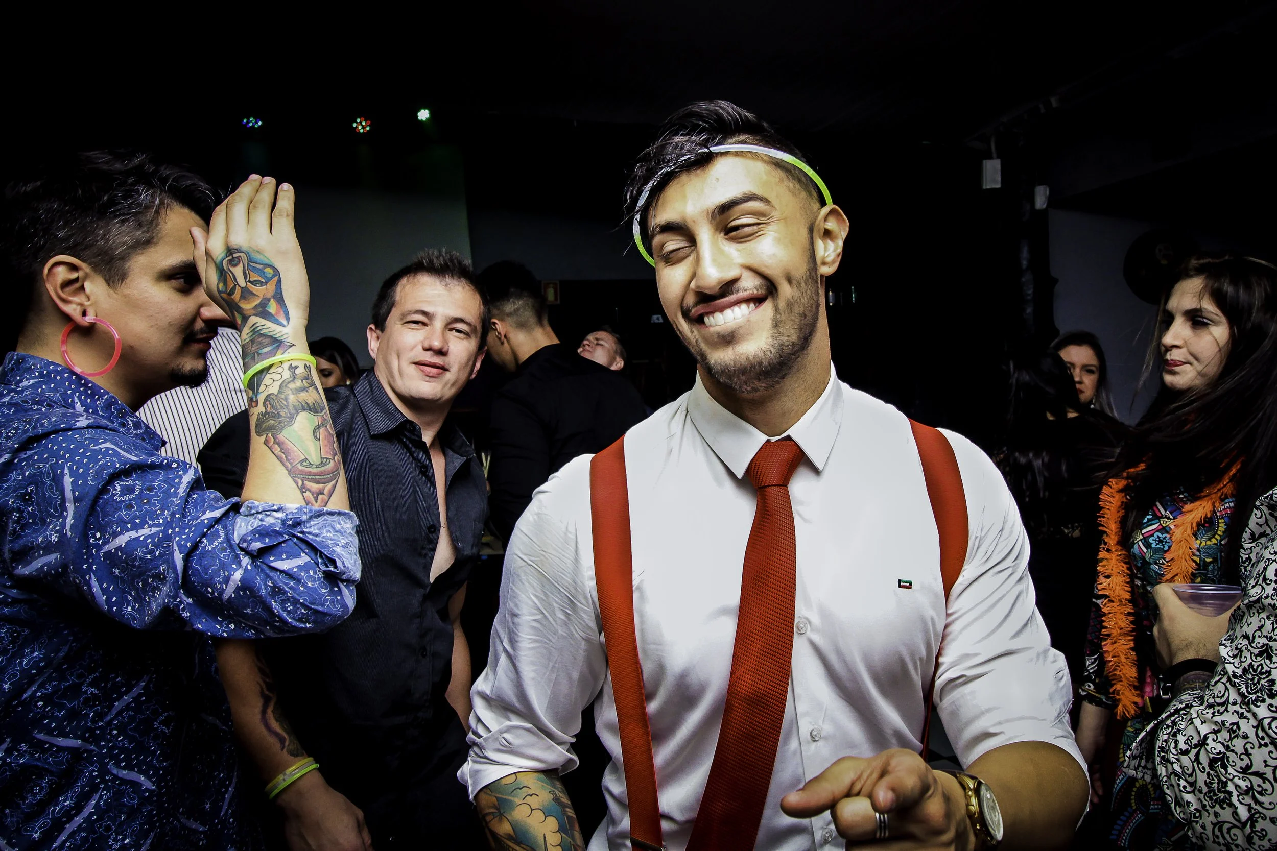 Homem sorridente com camisa branca, gravata vermelha, suspensórios vermelhos, e tatuagens, curtindo uma festa com várias pessoas ao fundo.