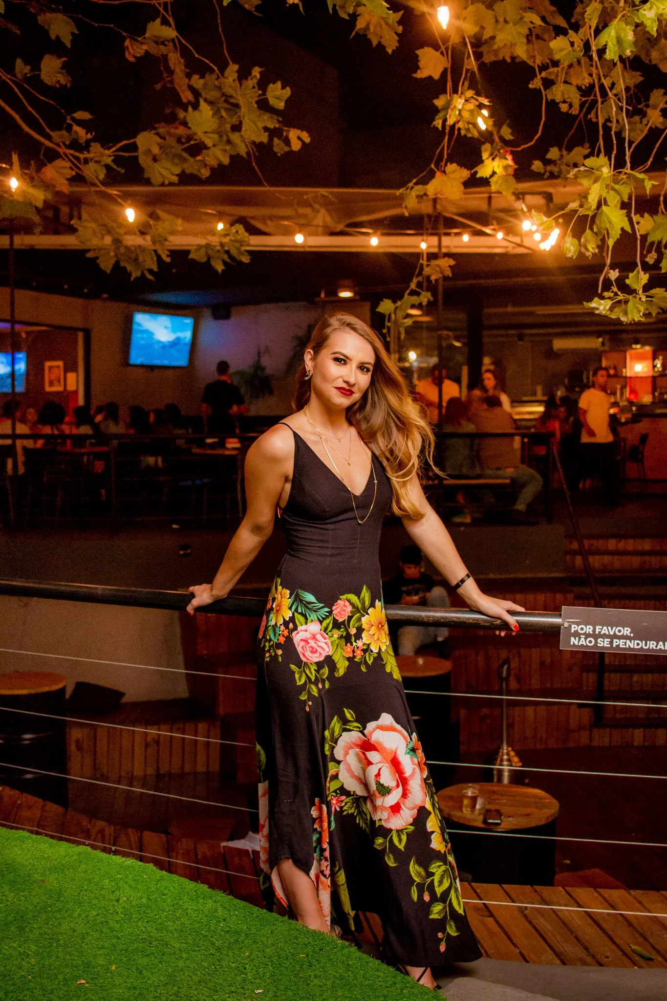 Mulher com vestido preto com estampa de flores sentada em um corrimão, em um ambiente de bar ou restaurante, com iluminação amarelada