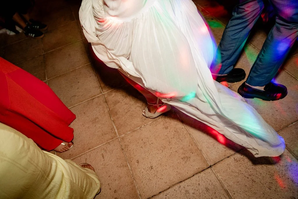 Pessoas em uma festa, com destaque para uma pessoa de vestido branco de cauda longa na pista de dança com iluminação de luzes de discoteca.