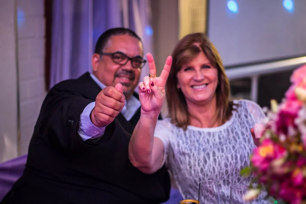 Homem e mulher sorrindo, fazendo o sinal de paz com as mãos, em uma festa ou celebração, com flores e taças na mesa.