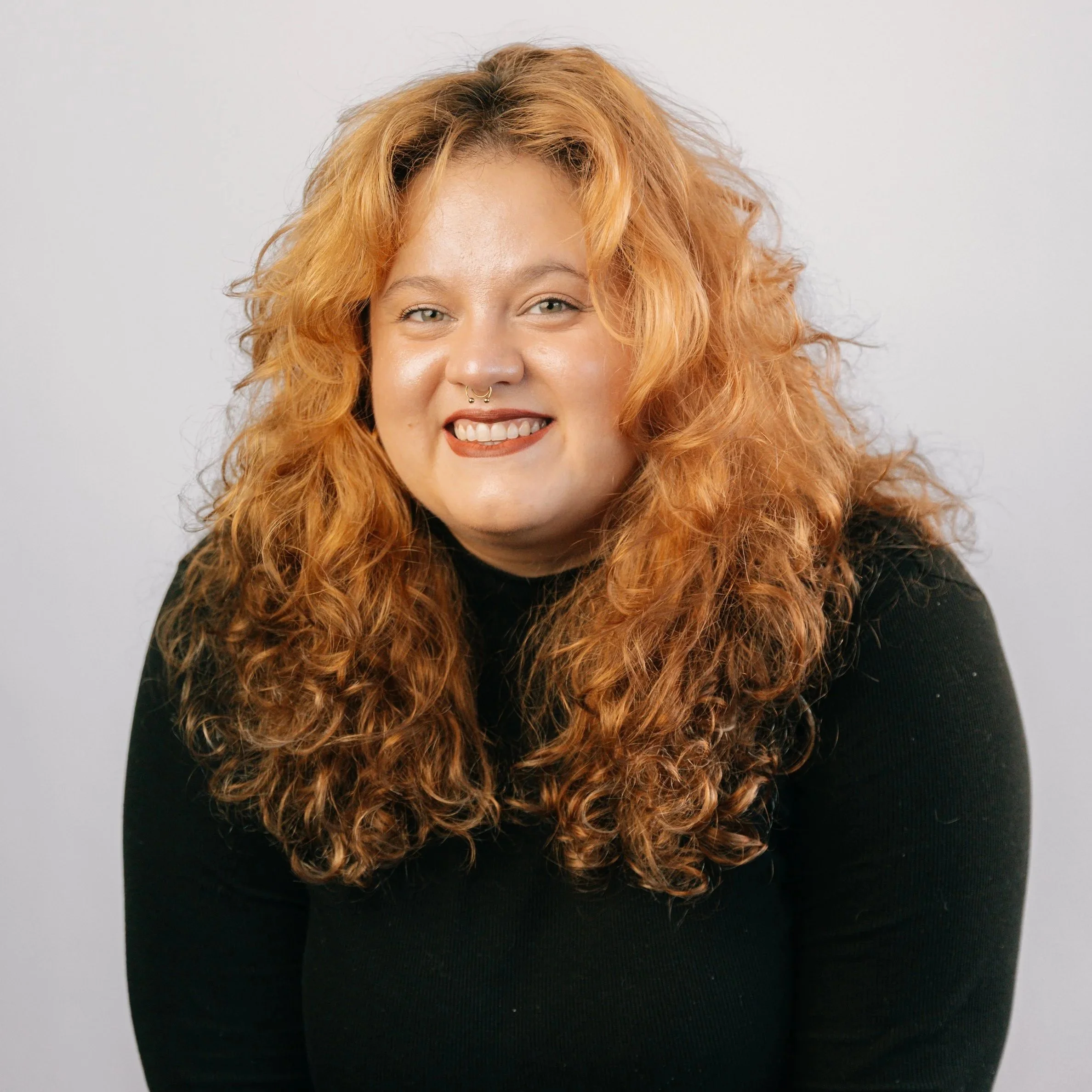 Mulher com cabelo ruivo cacheado, sorriso, usando maquiagem e um septo piercing, vestindo blusa preta contra fundo branco.