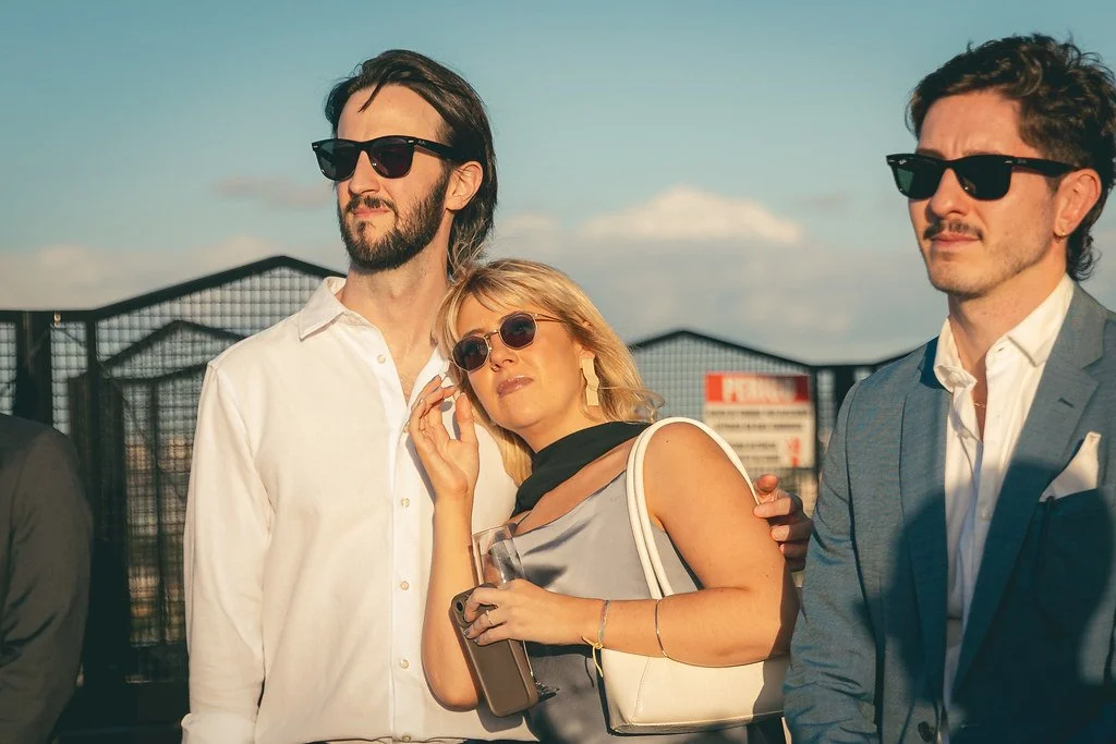 Três pessoas com óculos de sol, duas homens e uma mulher, posando ao ar livre durante o dia, com céu nublado, fundo de grade e sinalizações.