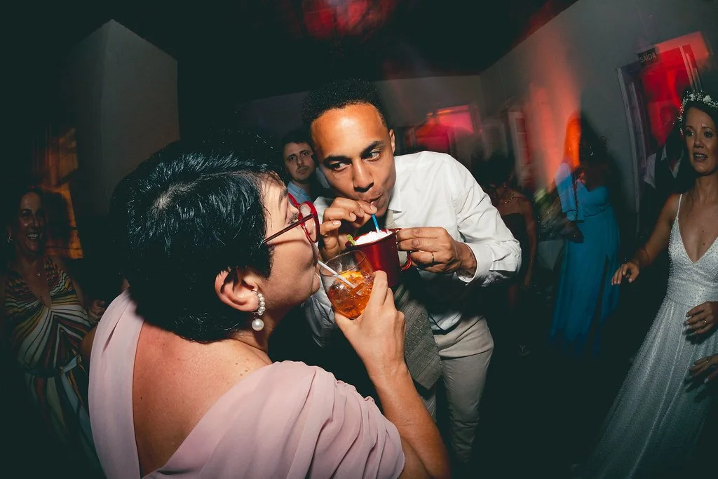 Pessoas celebrando em uma festa, com destaque para um homem e uma mulher que estão trocando bebidas, com outras pessoas ao fundo dançando e sorrindo