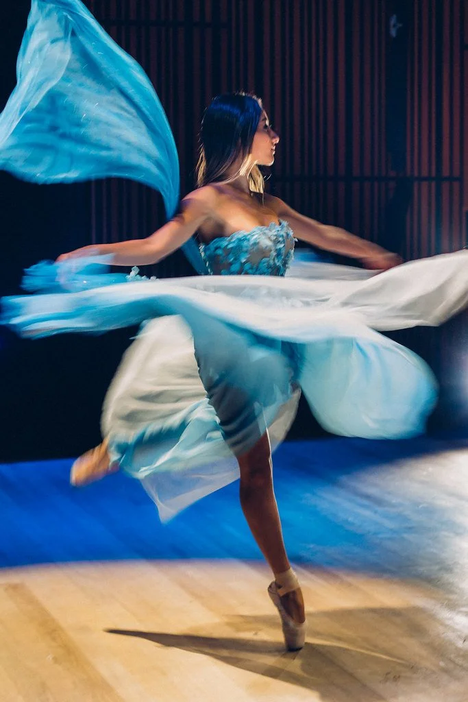 Bailarina em vestido azul fluido, dançando em palco de madeira com fundo escuro.
