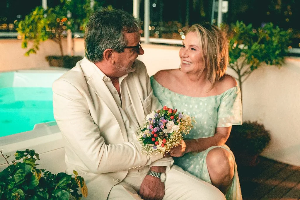 Um homem mais velho de óculos, vestindo um terno claro, segura um buquê de flores e sorri para uma mulher mais jovem, que está vestida com um vestido de ombro de cor verde claro, em um ambiente ao ar livre à noite, próximo a uma piscina