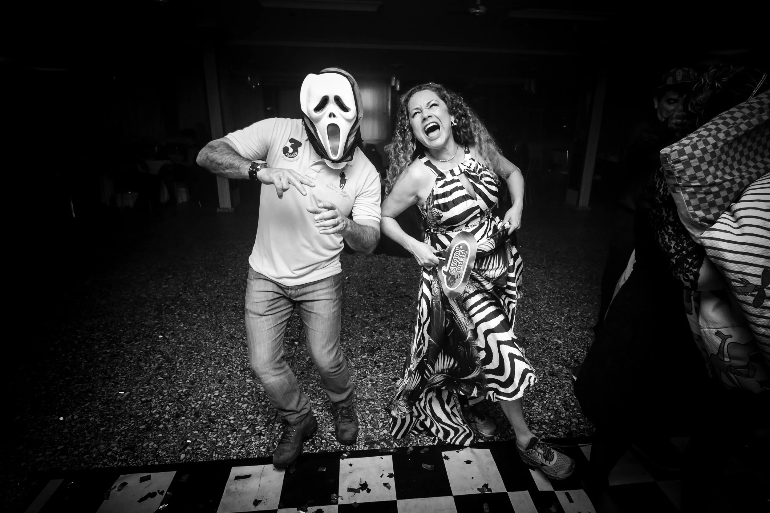 Pessoas se divertindo em uma festa; uma pessoa usando máscara de fantasma do filme 'Scream' e uma mulher com cabelo cacheado, vestindo vestido com estampa de zebra, rindo e posando ao lado dela.