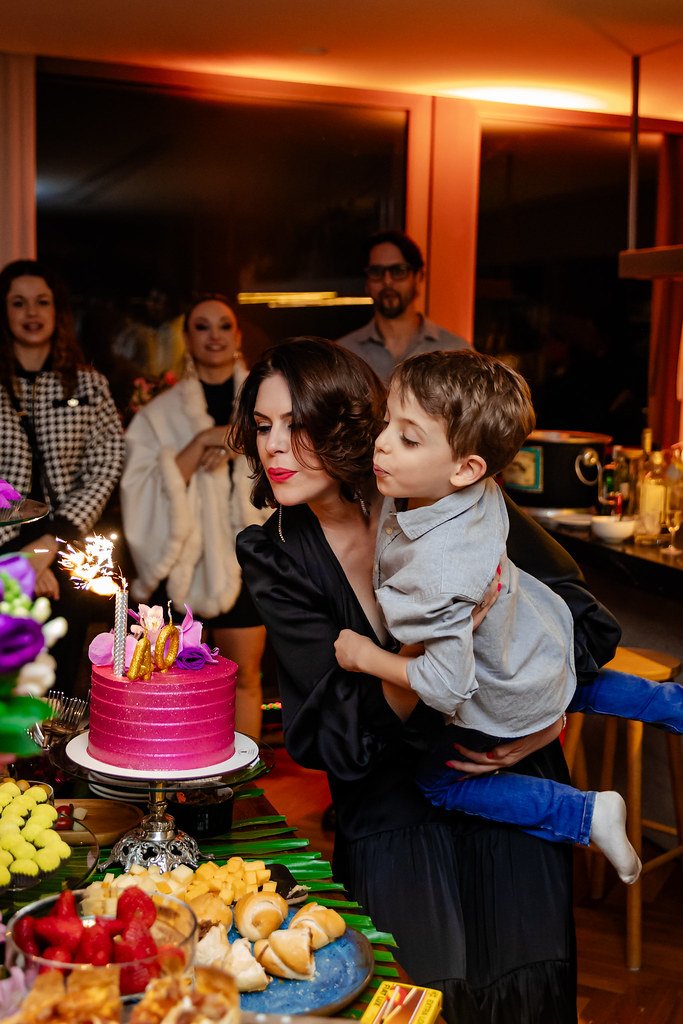 Mulher segurando menino perto de bolo de aniversário rosa com vela de vela, rodeada por pessoas em comemoração