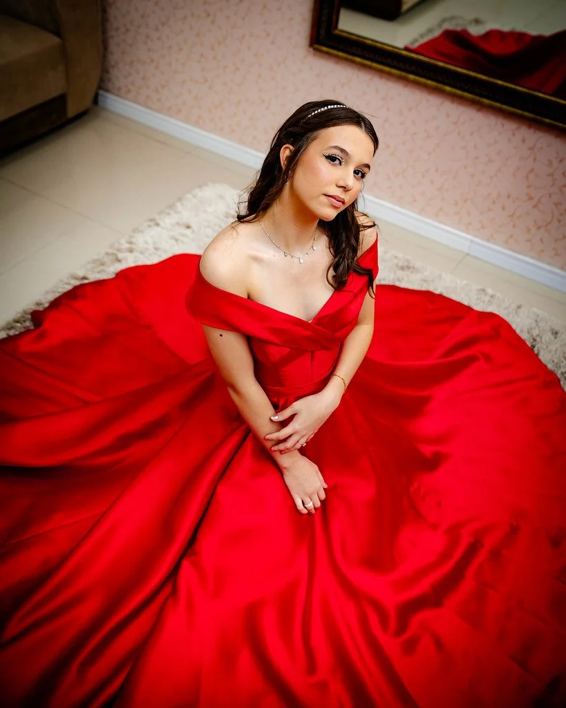 Mulher jovem com vestido vermelho longo, sentada no chão com o vestido espalhado ao redor, no interior de uma sala com parede de papel de parede e espelho na parede.