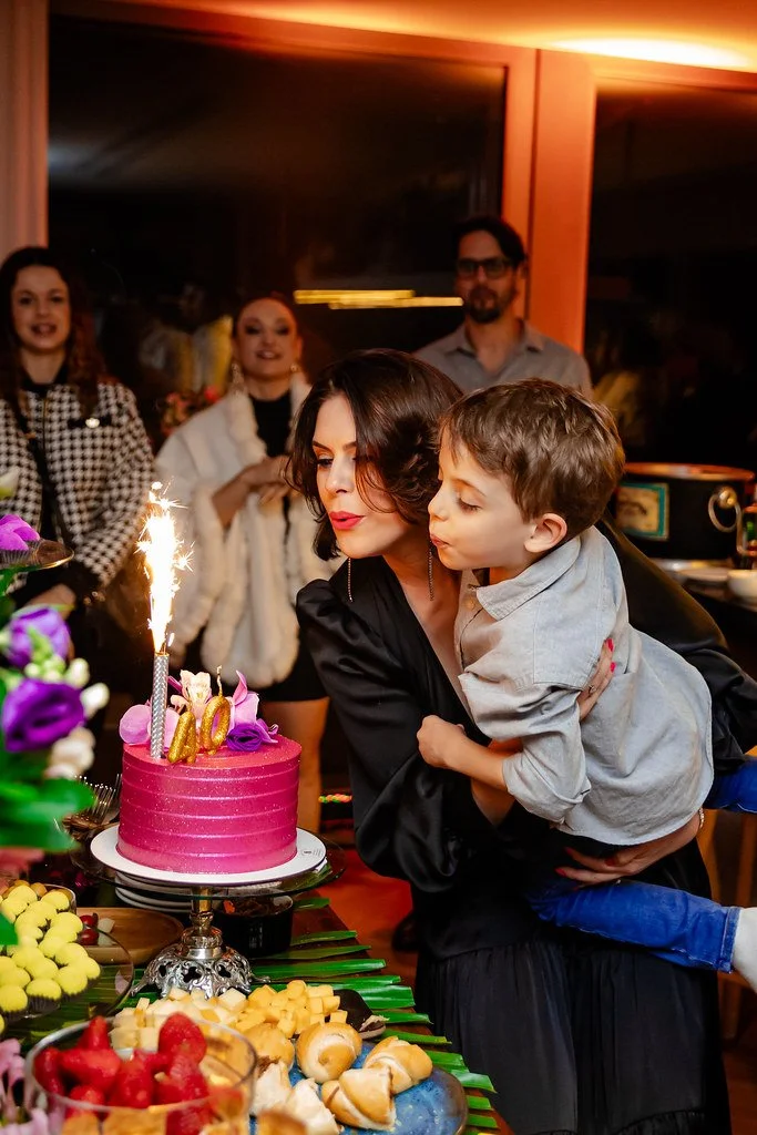 Mulher segurando uma criança na frente de um bolo de aniversário rosa com vela e decoração, com várias pessoas ao fundo em uma celebração.