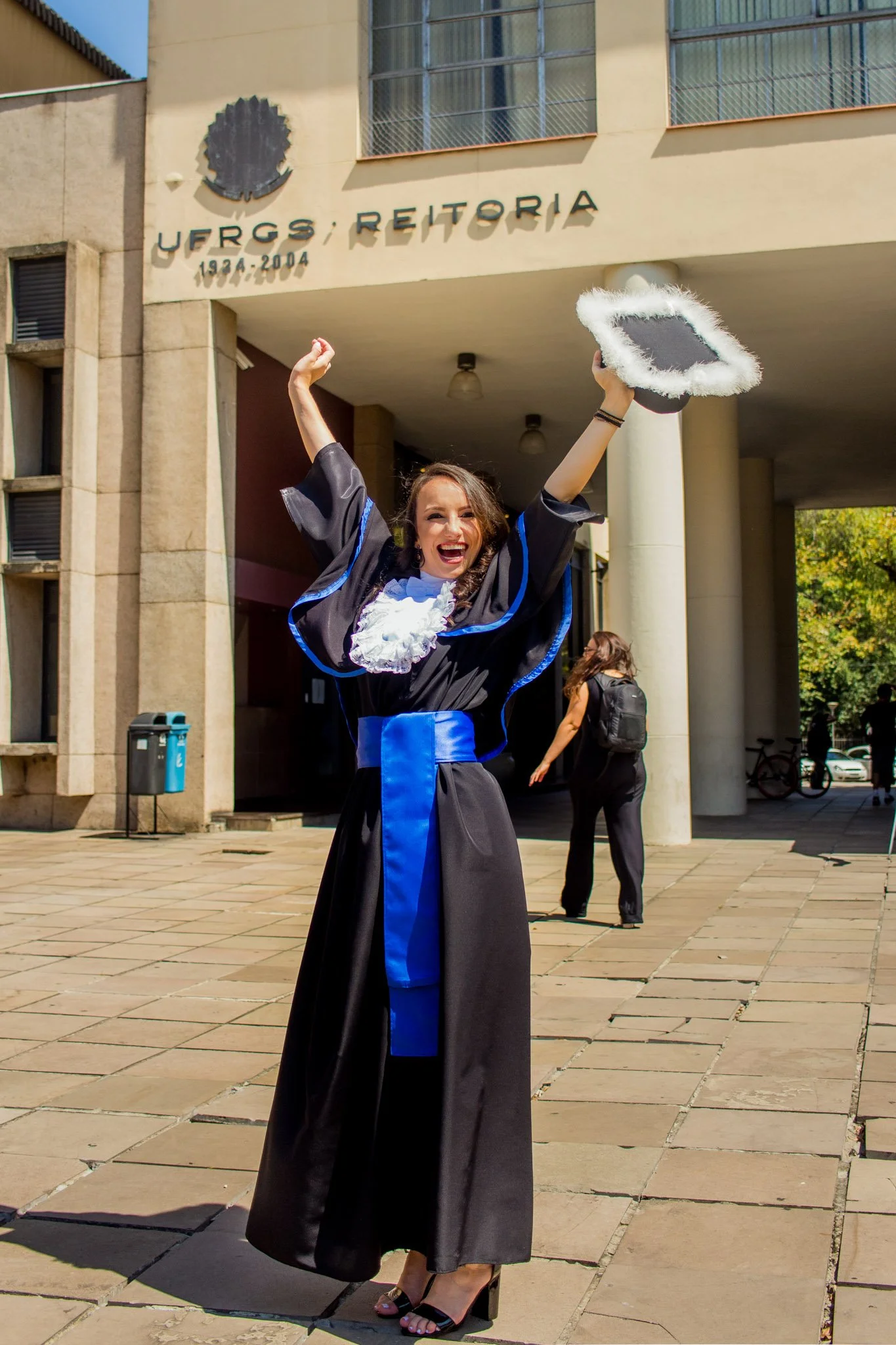 Mulher vestida de formado universitário celebrando com chapéu de formatura na frente da universidade.