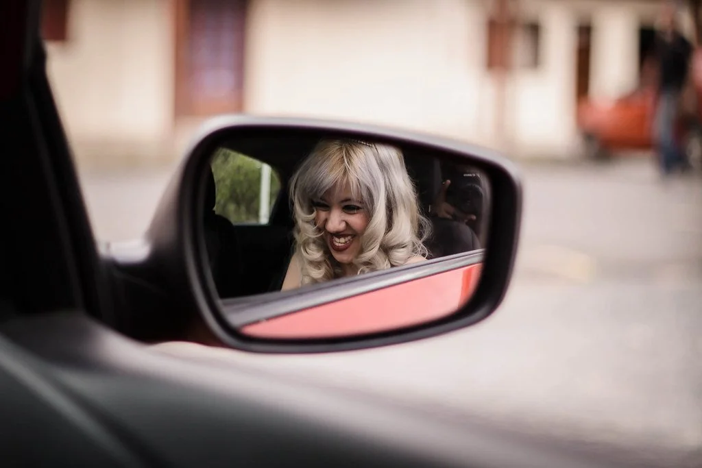 Pessoa com cabelo loiro cacheado sorrindo ao olhar a reflexão na janela lateral de um carro.