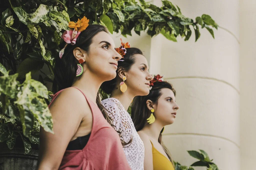 Três mulheres jovens de perfil, usando roupas coloridas e acessórios de flores, observando ao lado de uma parede com vegetação.