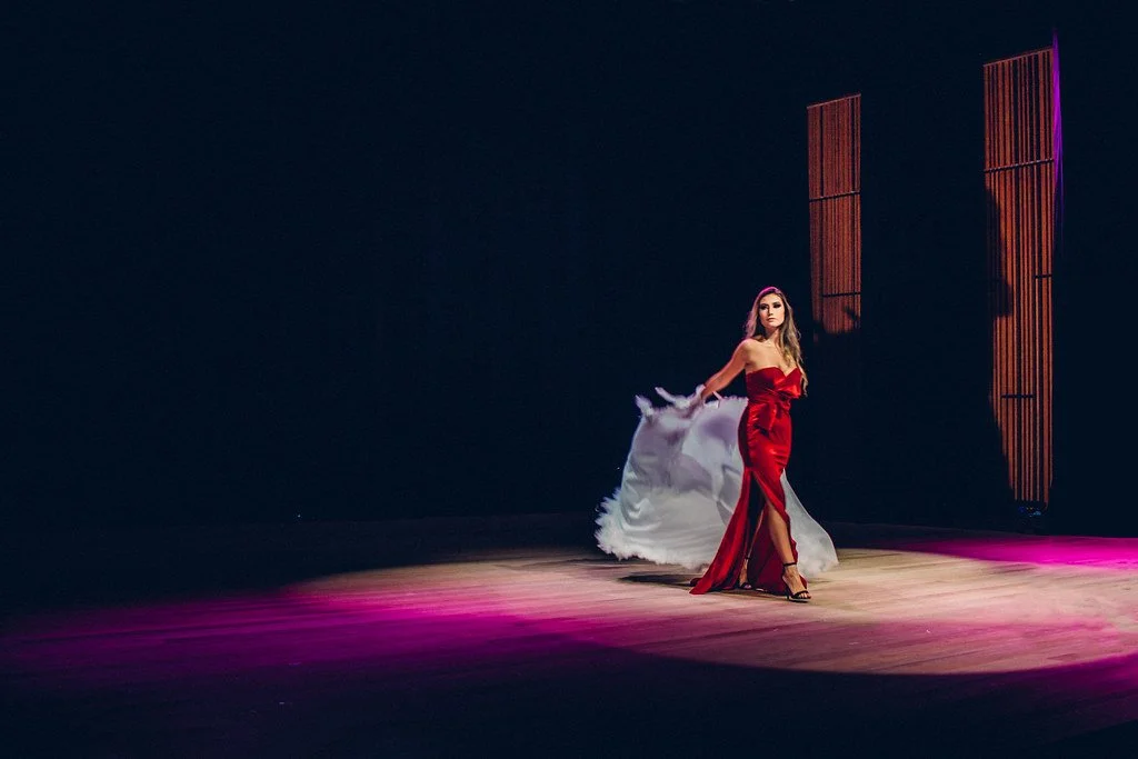 Mulher em vestido longo vermelho caminhando no palco com iluminação rosa e fundo escuro.