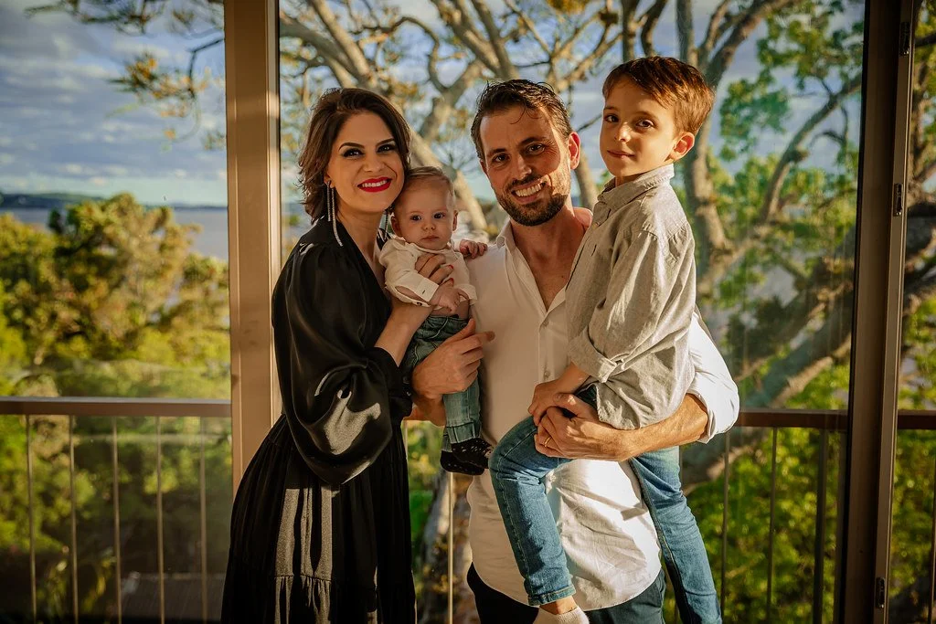 Família sorridente em ambiente ao ar livre, com árvores e céu ao fundo.