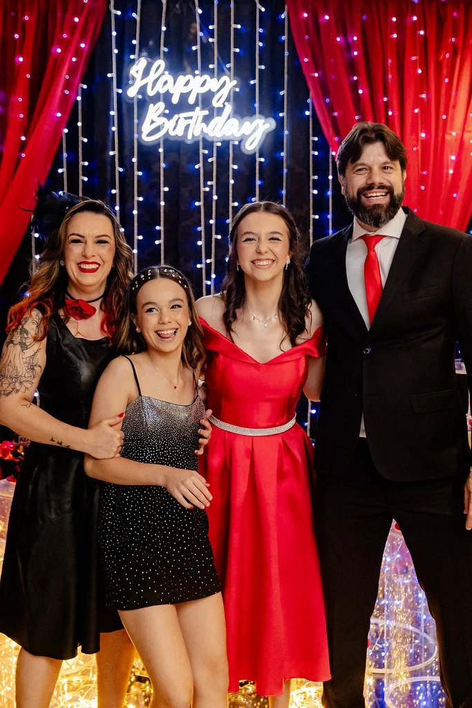 Família celebrando aniversário de cabelo preto, roupas de festa, na decoração com luzes e painel com 'Happy Birthday' ao fundo.