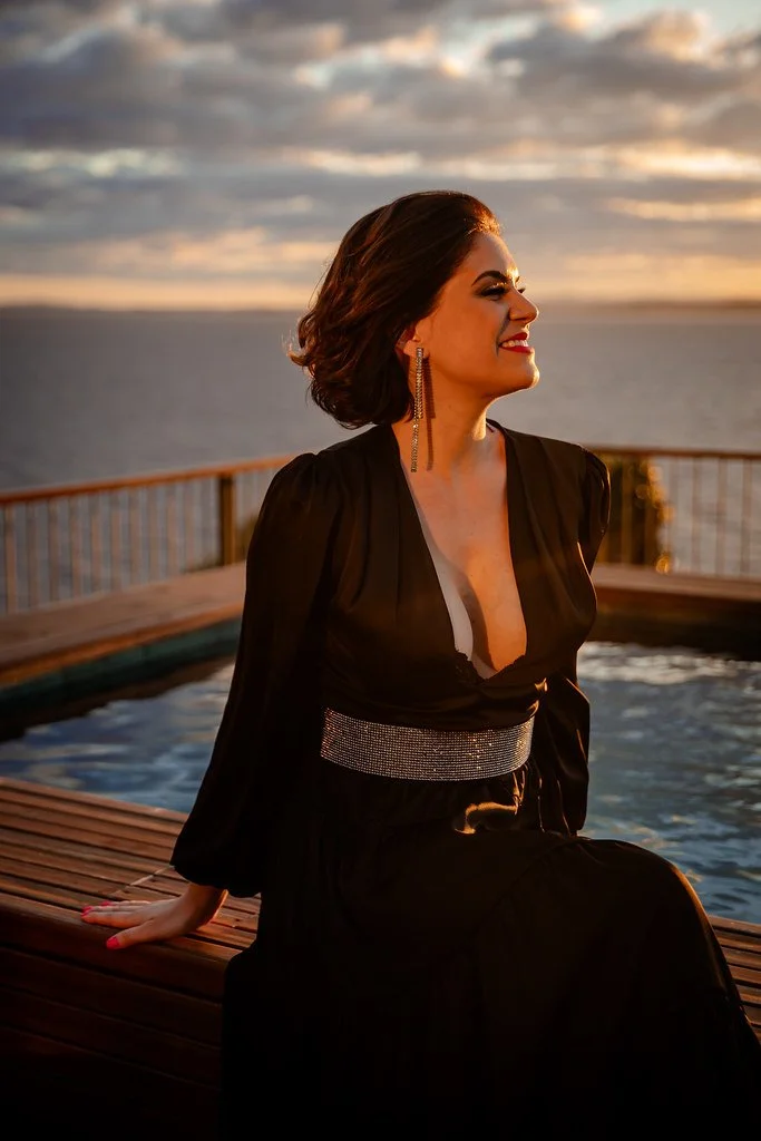 Mulher sorridente com cabelo curto e vestido preto, sentada ao lado da piscina com vista para o mar ao pôr do sol.