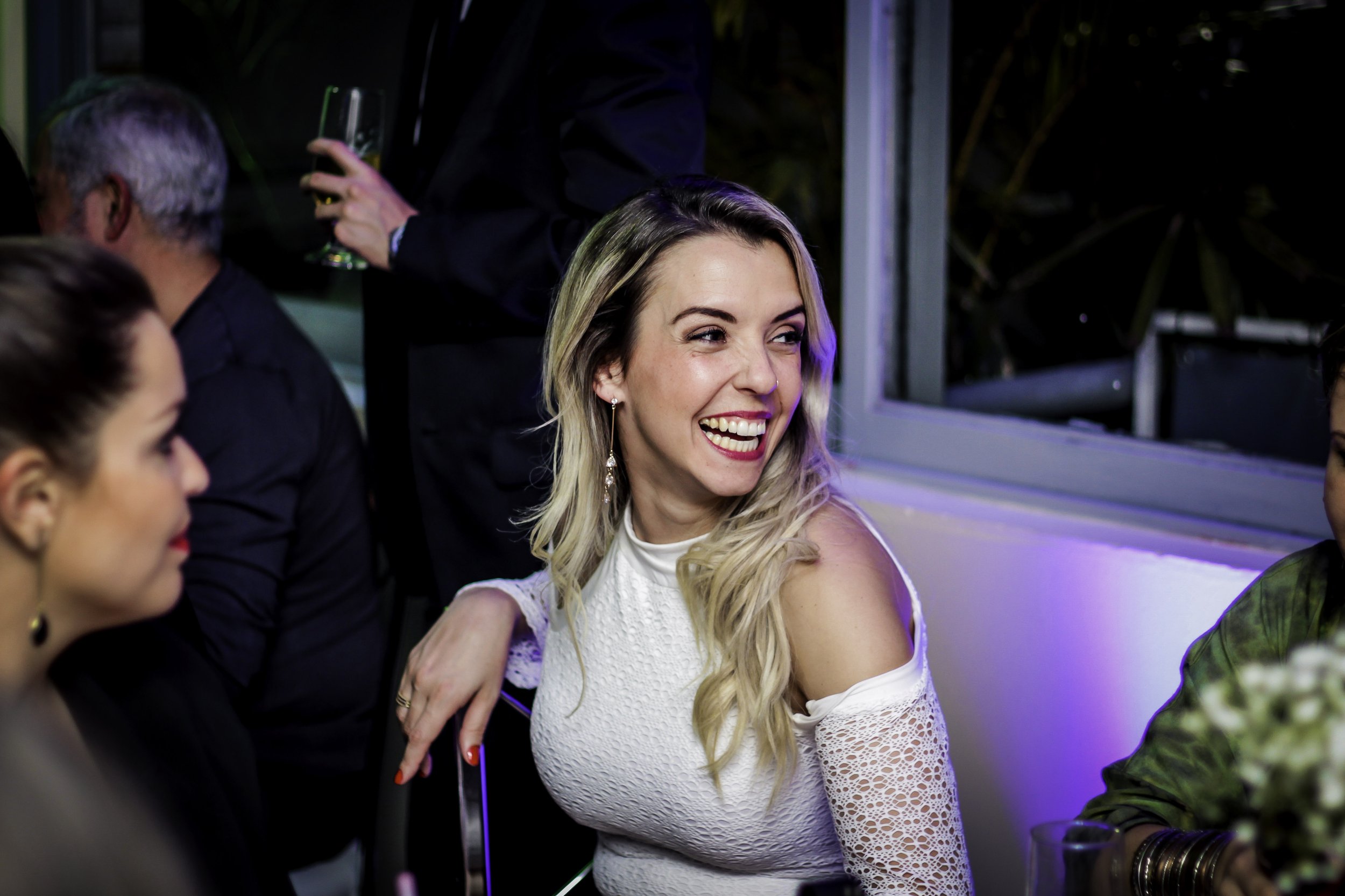 Mulher sorrindo em festa, usando vestido branco com detalhes de renda, com cabelo loiro ondulado, ao lado de outras pessoas em ambiente festivo com luzes coloridas.