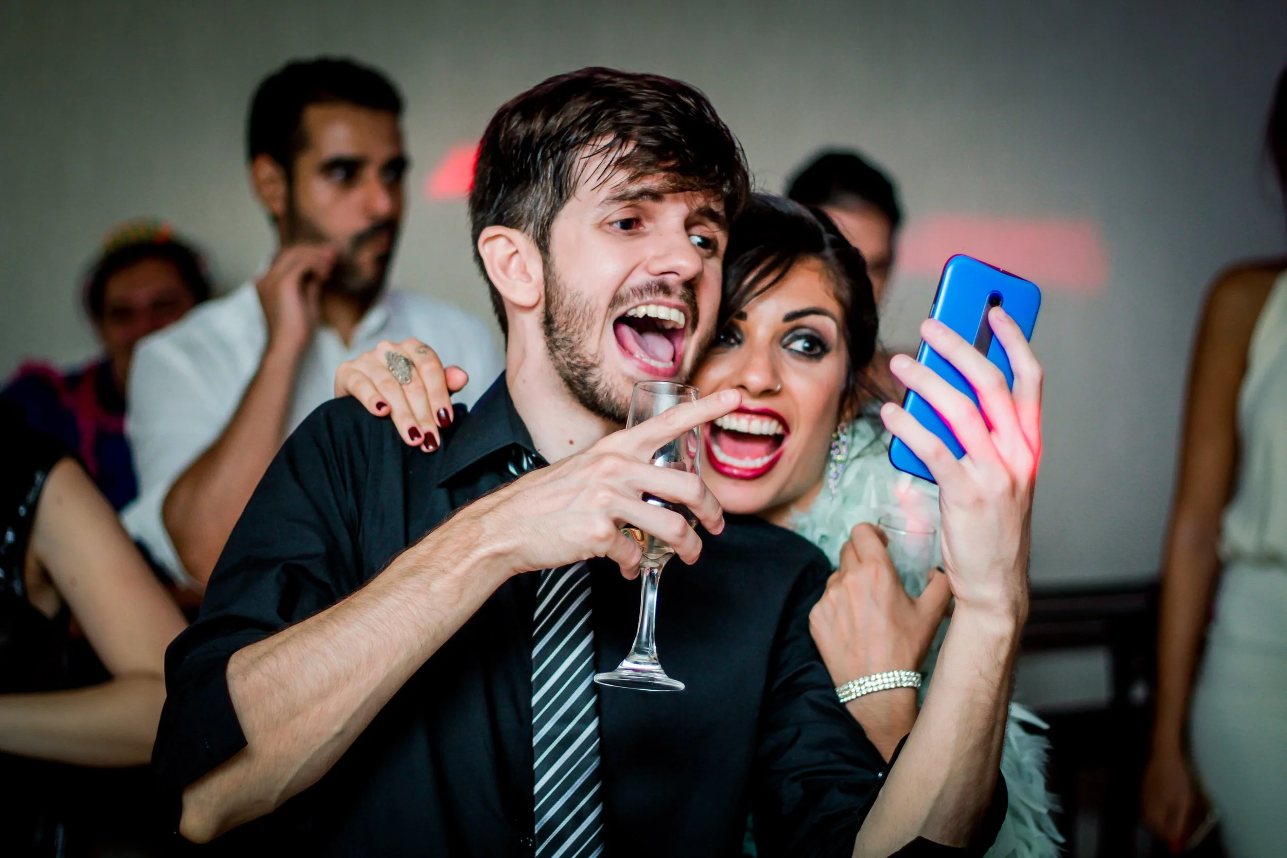 Pessoas felizes tirando foto em uma festa, com um homem de cabelo castanho, barba e vestido social preto e uma mulher com cabelo escuro, maquiagem marcante e roupa brilhante. Ao fundo, outras pessoas observando, em um ambiente festivo.
