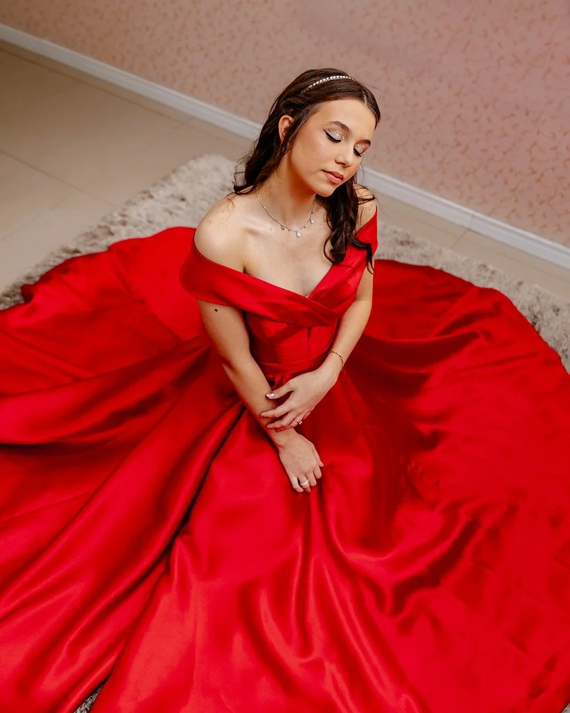 Mulher com vestido vermelho longo, sentada de cabeça inclinada, com cabelo escuro e acessório na cabeça, em ambiente interno.