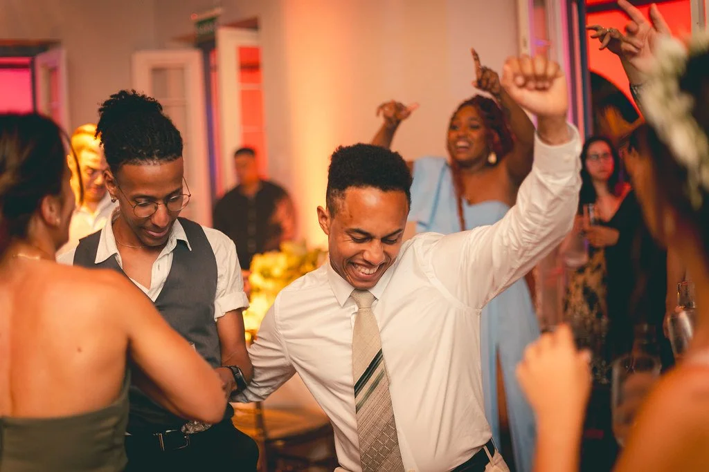 Pessoas dançando e se divertindo em uma festa, com um homem sorridente e braços levantados no centro.