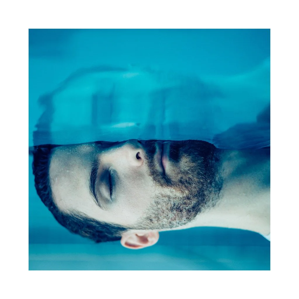 Homem com barba e cabelo molhados, de olhos fechados, com expressão tranquila, sob água azul.