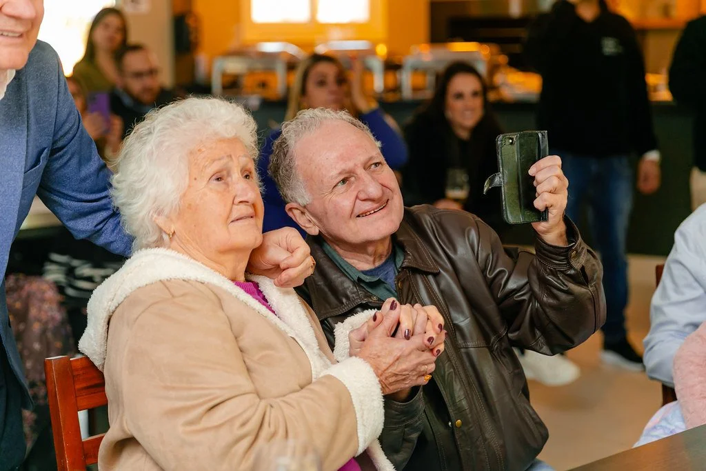 Um casal idoso sentado junto, a mulher com cabelo branco e vestindo um casaco bege, e o homem com cabelo cinza, usando jaqueta de couro, sorrindo enquanto tira uma foto com o celular. Ao fundo, várias pessoas assistindo e sorrindo.