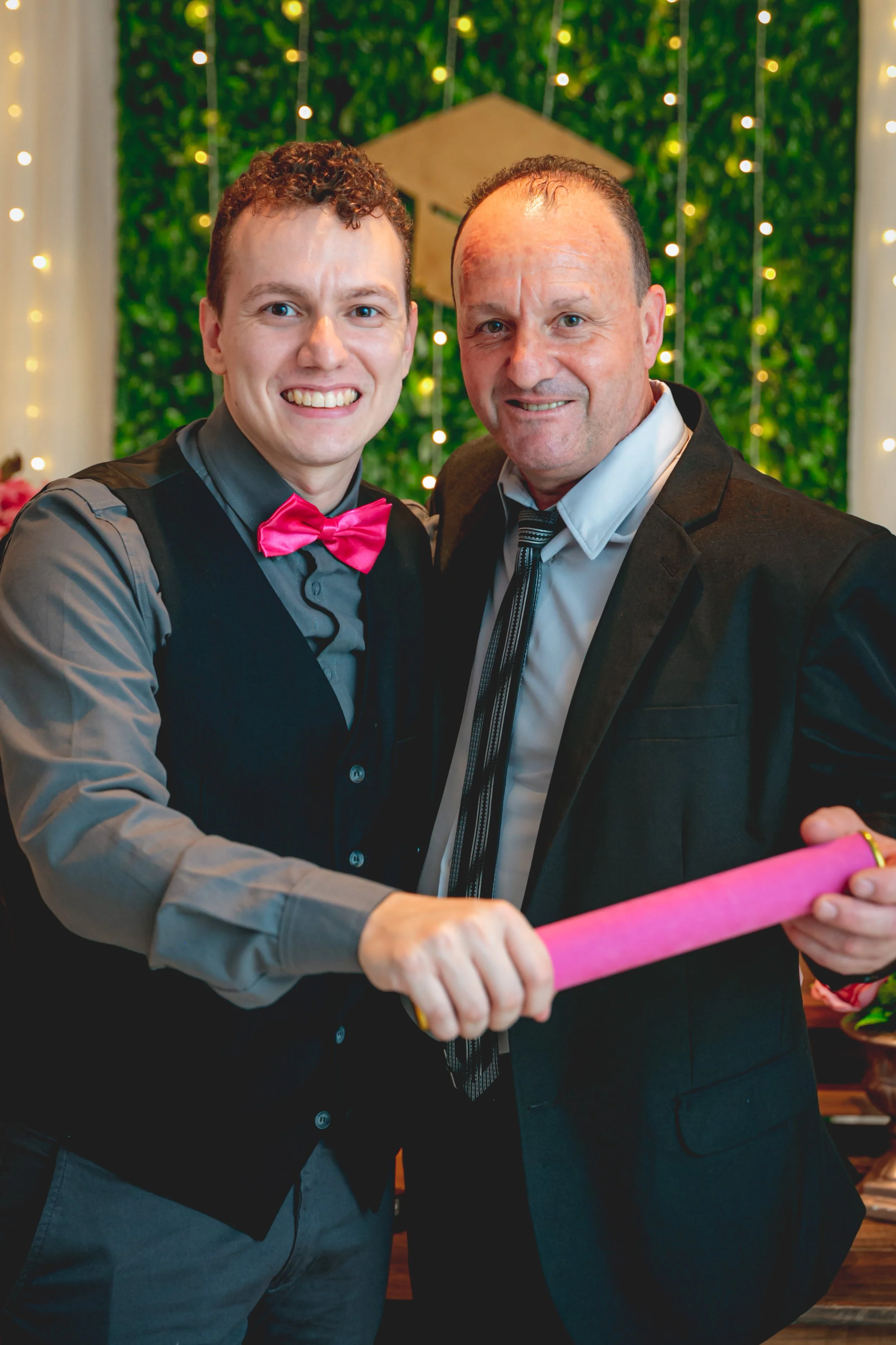 Duas pessoas em traje formal celebrando, uma delas segurando um rojão, com fundo decorativo de luzes e plantas.