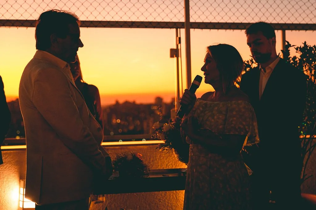 Pessoa falando ao microfone durante pôr do sol em uma cobertura com outras pessoas ao fundo.