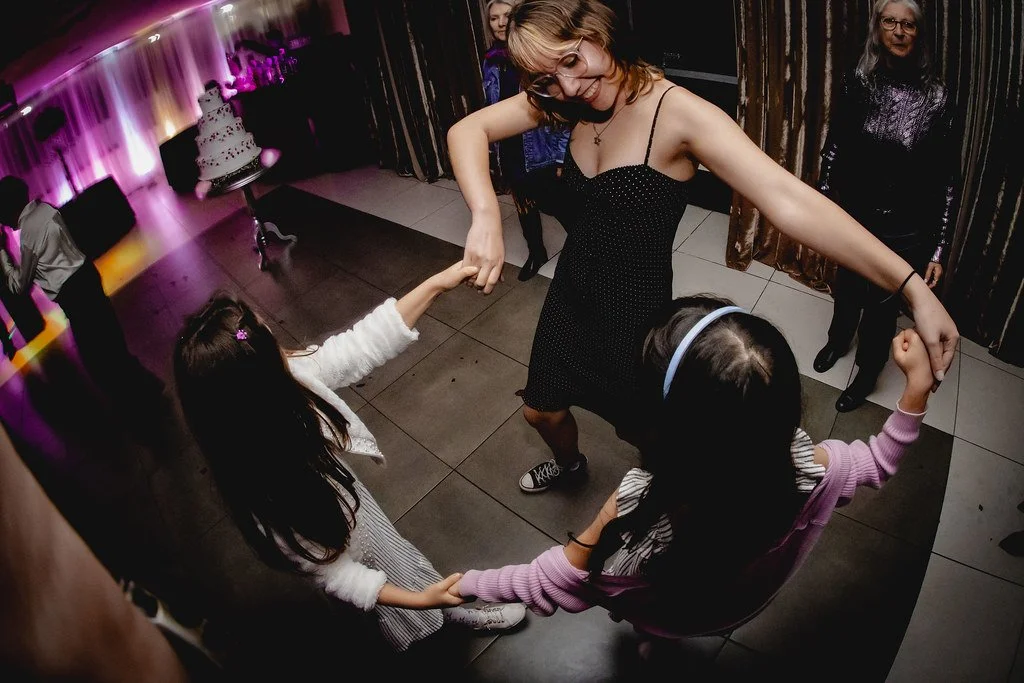 Mulher dançando com duas crianças sentadas, segurando as mãos enquanto giram em cima de uma pista de discoteca com iluminação colorida em um evento social.