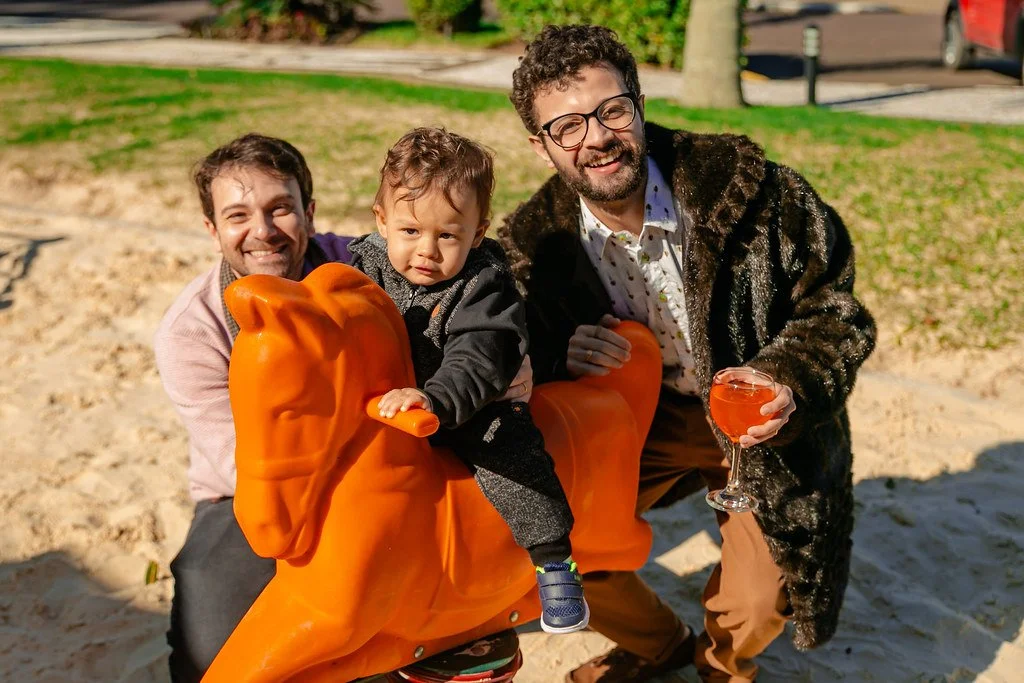 Três pessoas, dois adultos e uma criança, posando com uma escultura de cavalo alaranjada em um parque ao ar livre. Os adultos seguram uma taça de bebida de cor laranja, e todos parecem felizes.