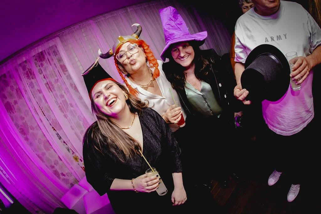 Mulheres em uma festa usando fantasias de bruxa, cavaleiro vikings e chapéu de mágico, sorrindo e se divertindo.