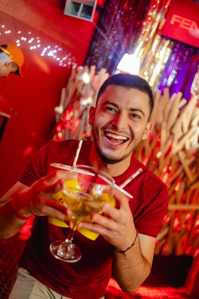Homem sorridente segurando uma bebida em um ambiente decorado com luzes coloridas e elementos de festa.