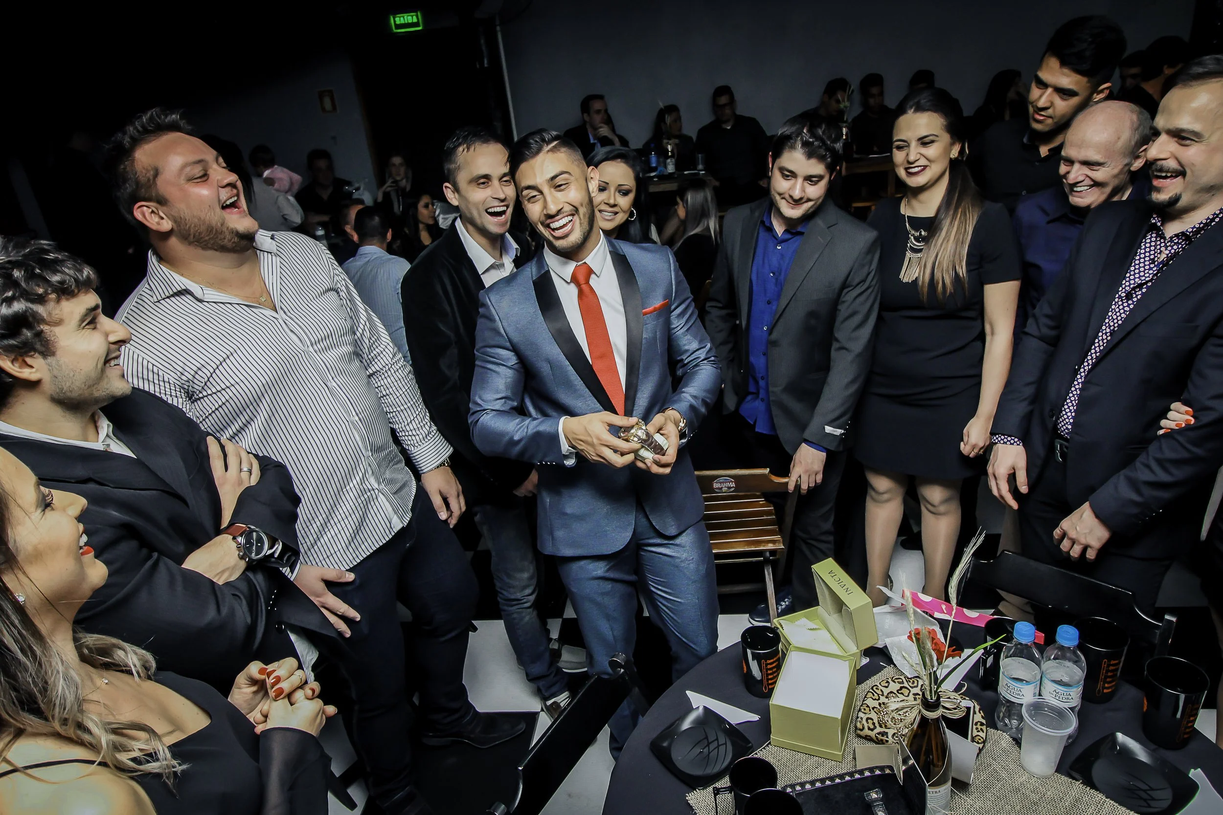 Homens e mulheres em um evento social, com um homem no centro segurando uma quantia de dinheiro, todos sorrindo e conversando em um ambiente interno.