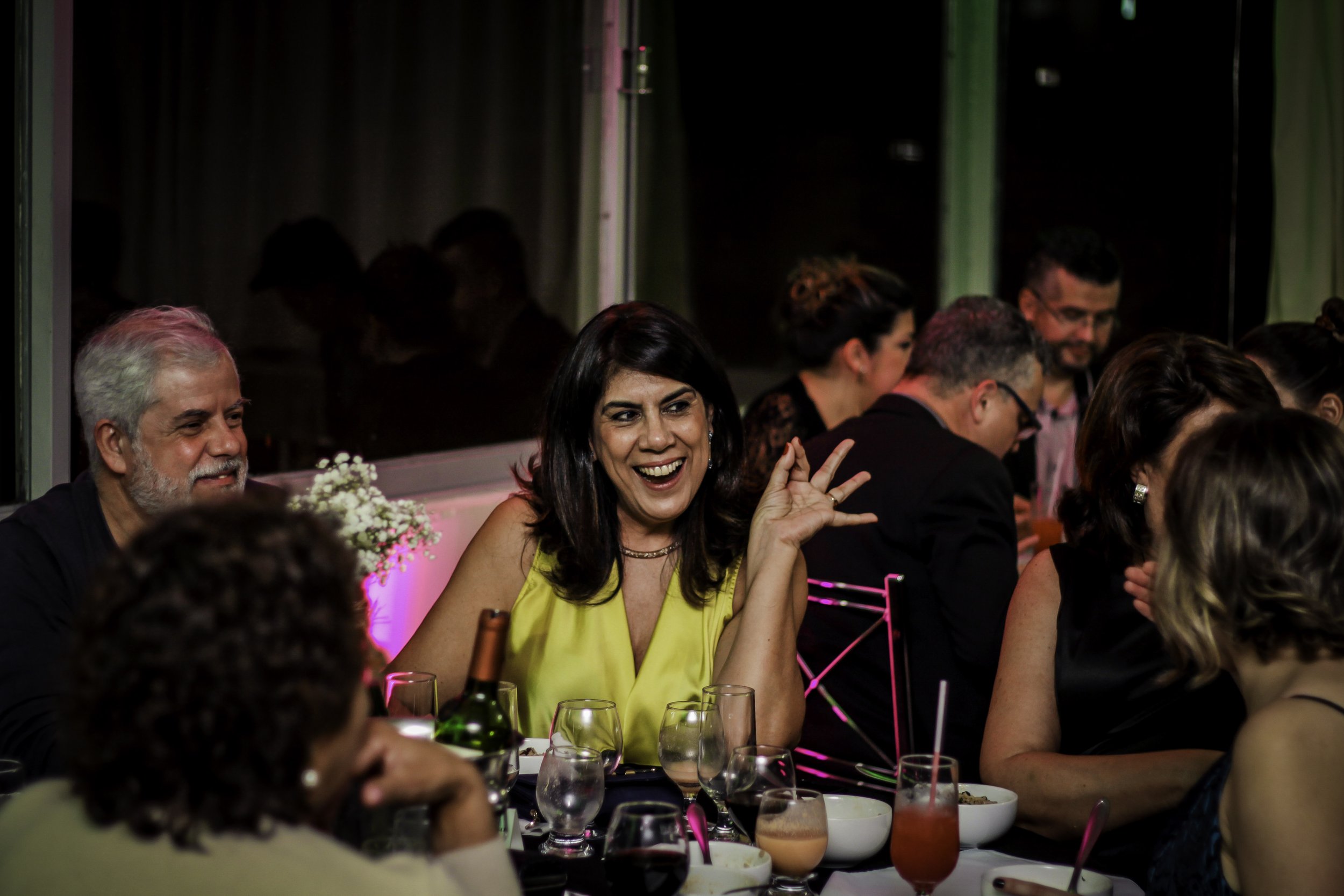 Grupo de pessoas comemando em festa, com uma mulher sorridente de vestido amarelo no centro.