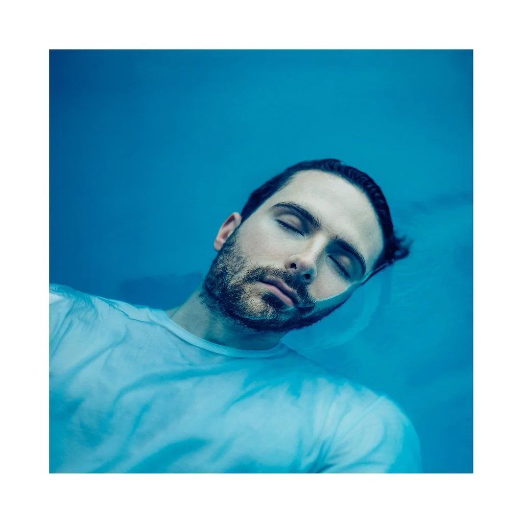 Homem de cabelo escuro e barba, com expressão tranquila, boiando de olhos fechados em água azul.