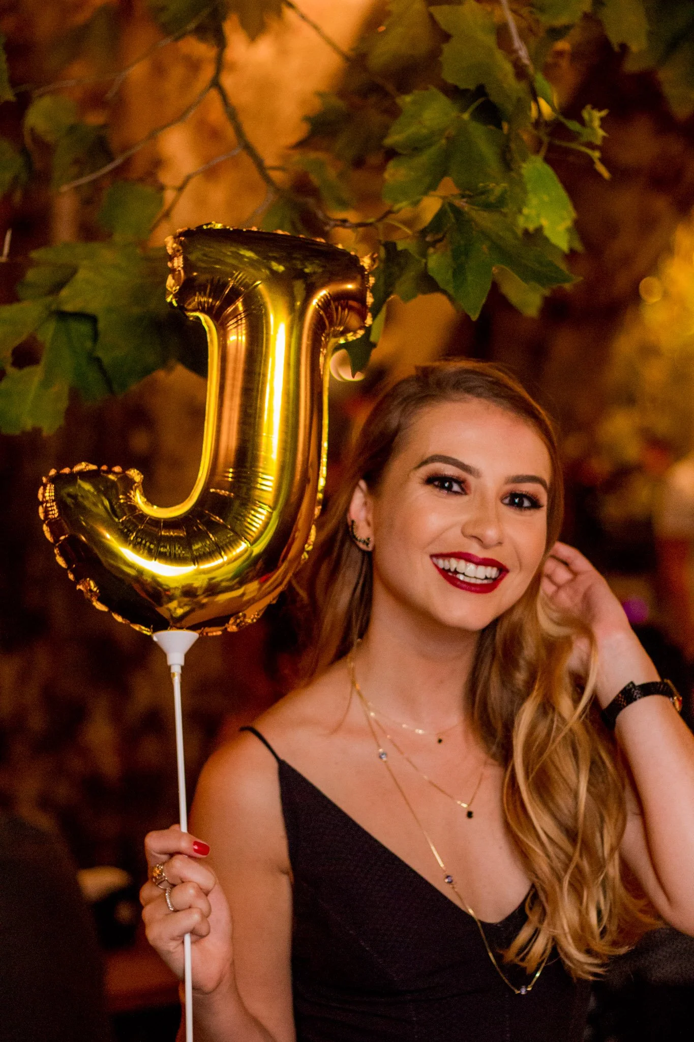 Mulher sorridente segurando um balão dourado em forma de letra J, em ambiente com iluminação quente e folhas verdes ao fundo.