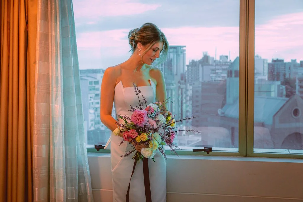Mulher de vestido branco segurando um buquê de flores com fundo de janela e cidade ao entardecer.