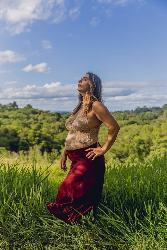 Mulher posando em campo com céu azul e nuvens ao fundo, usando top bege e saia longa vermelha.