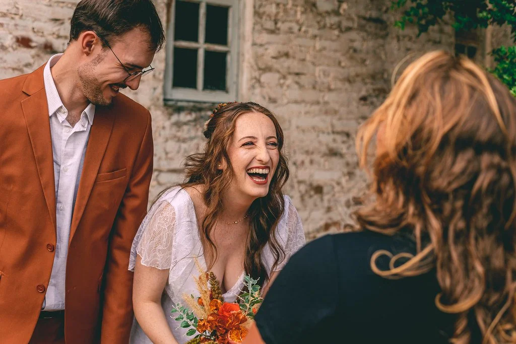 Mulher sorridente em casamento segurando um buquê de flores, ao lado de homem de blazer marrom, com duas pessoas ao fundo, em ambiente ao ar livre.