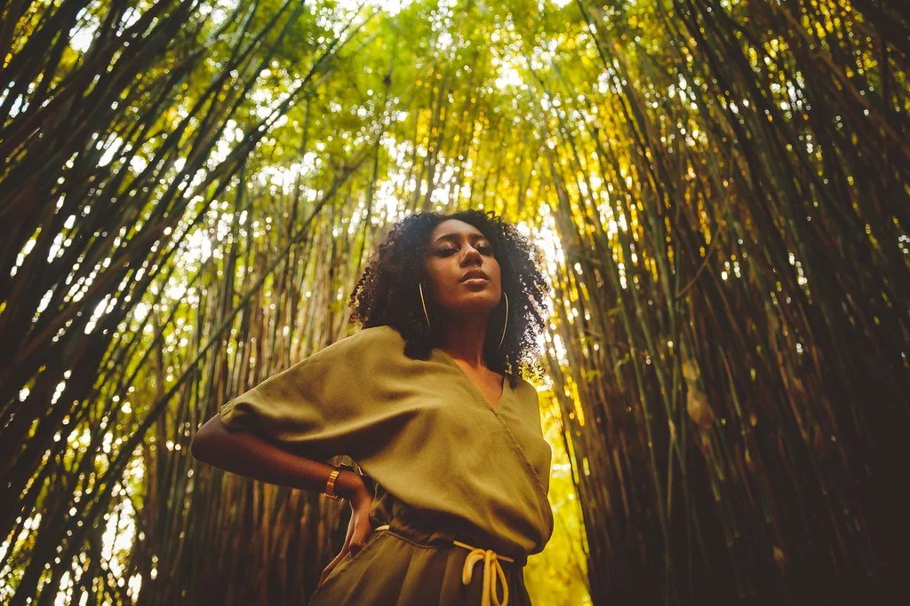 Mulher com cabelo cacheado em um bosque de bambu, vestindo uma blusa de manga curta e calça com cinto, com luz solar filtrando pelas folhas.