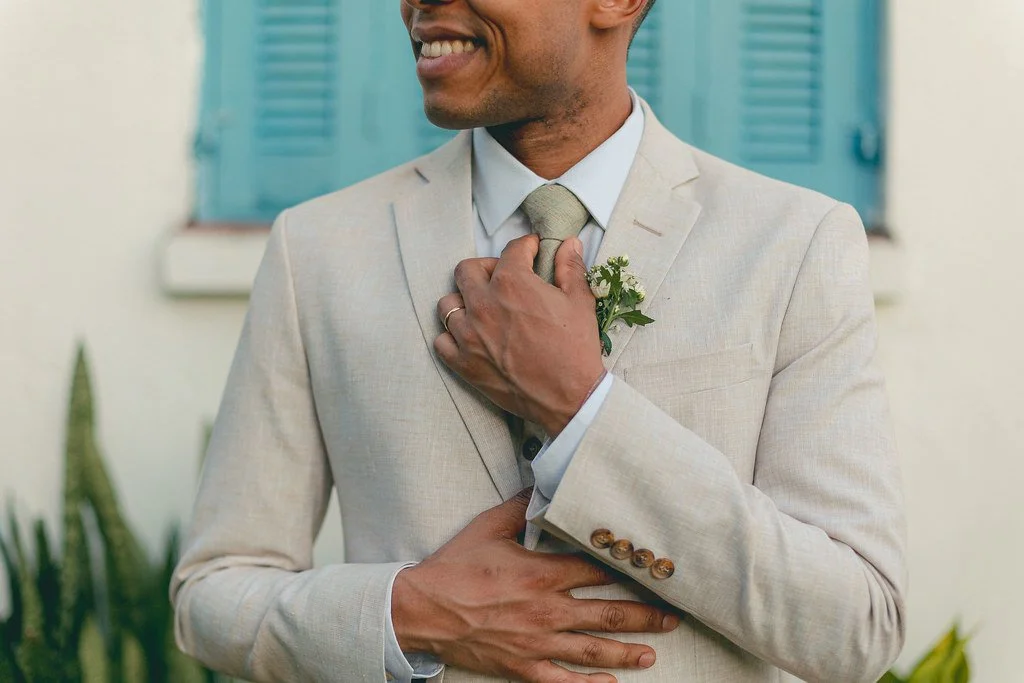 Homem sorridente vestido com terno claro, ajustando sua gravata, com uma pequena flor presa na lapela, em frente a uma parede com janela de persianas azuis.