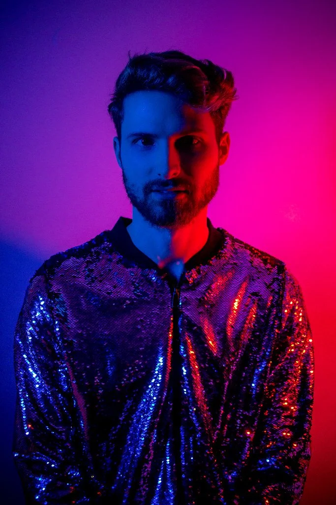 Homem com cabelo escuro, barba e jaqueta de paetês brilhantes com iluminação azul e rosa.