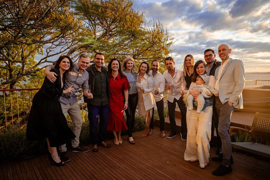 Grupo de pessoas felizes em uma reunião ao ar livre ao pôr do sol, com árvores ao fundo e uma varanda de madeira.