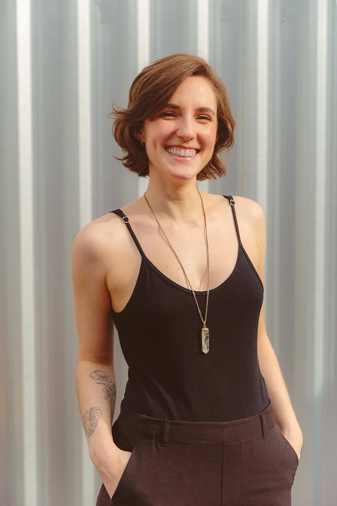 Mulher sorrindo, com cabelo curto castanho, vestindo blusa preta de alças finas, um colar com pingente e calças pretas, diante de um fundo de parede de metal.