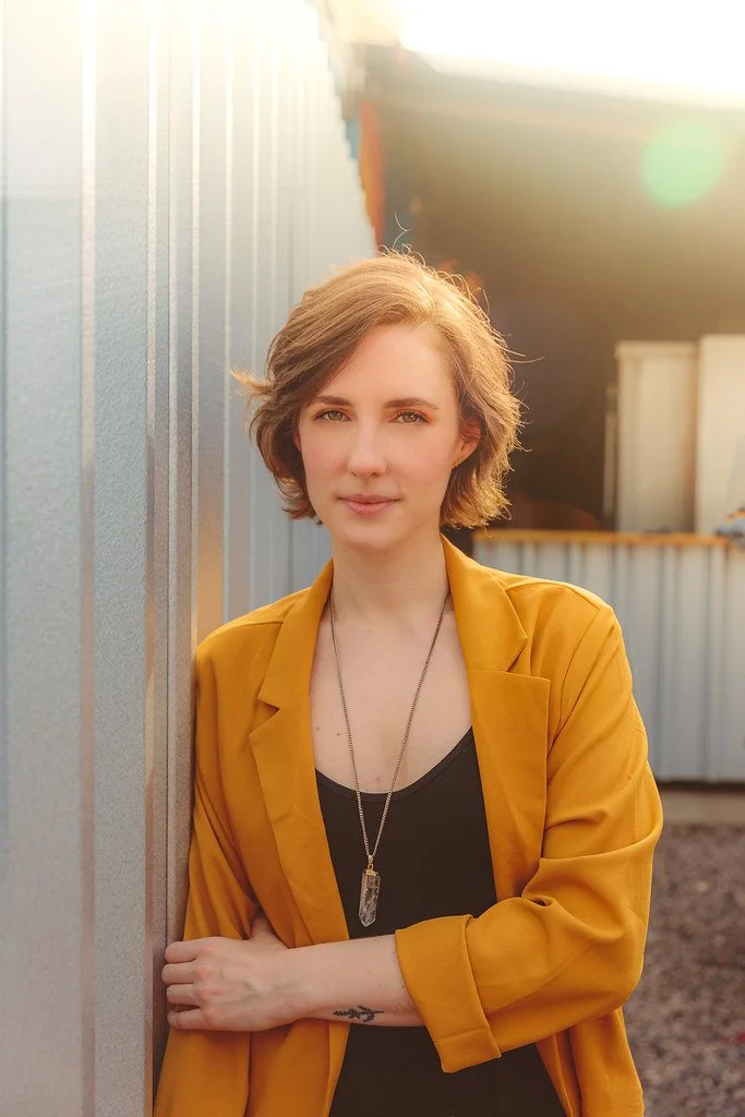 Retrato de uma mulher com cabelo curto e castanho claro, usando jaqueta amarela e camiseta preta, posando ao lado de uma parede metálica ao pôr do sol.